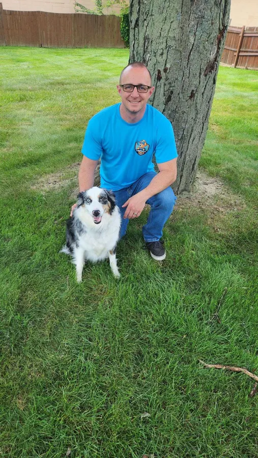 Landmine Patrol team member kneeling with an Australian shepherd in a fenced Akron-area yard
